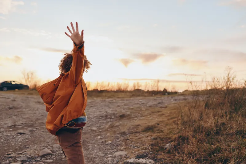 Woman lifestyle outdoor at sunset, walking on rural road, arms raised waving hands, wearing warm jacket, golden sky and soft clouds, peaceful mood, natural landscape, evening