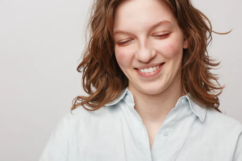 Smiling young woman with curly hair wearing white shirt looks down with closed eyes. Studio portrait on light background. Concept of happiness, natural beauty, and casual lifestyle.