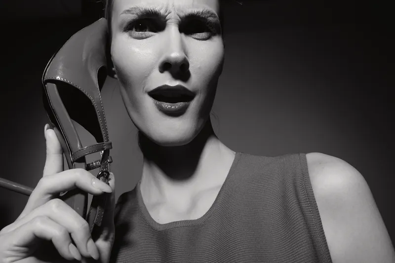 Close-up of a fair skinned young woman holding a high heel shoe to her ear with a confused expression on her face in a studio with dramatic lighting.