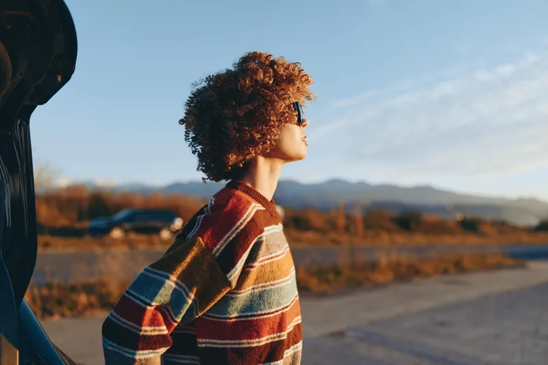 Woman lifestyle outdoors portrait side profile with curly hair, striped poncho, standing on road near mountains, open landscape at dusk, calm thinking mood, natural candid moment