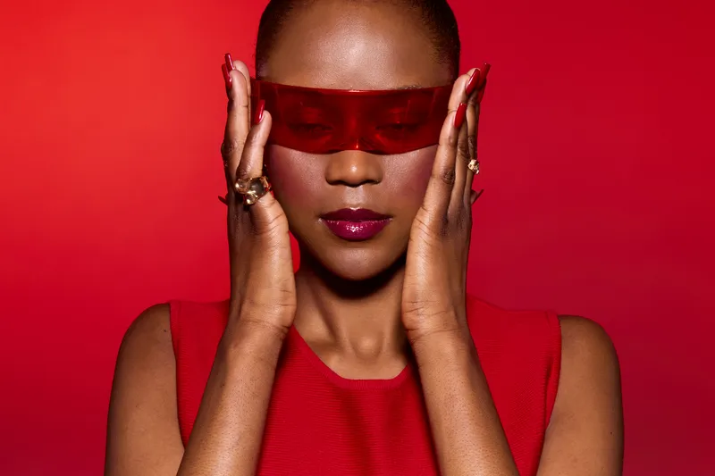 Portrait of a confident black woman with dark skin wearing red futuristic glasses and a red sleeveless top. The red background enhances the bold and stylish concept of fashion and beauty.