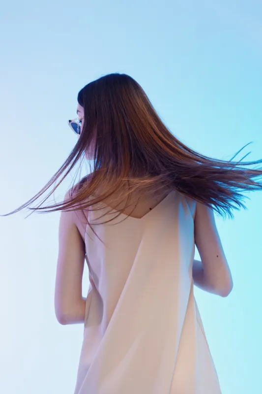Young woman with long hair wearing a beige sleeveless dress turns her head in a studio with blue lighting. Concept of beauty, fashion, and elegance in portrait photography.