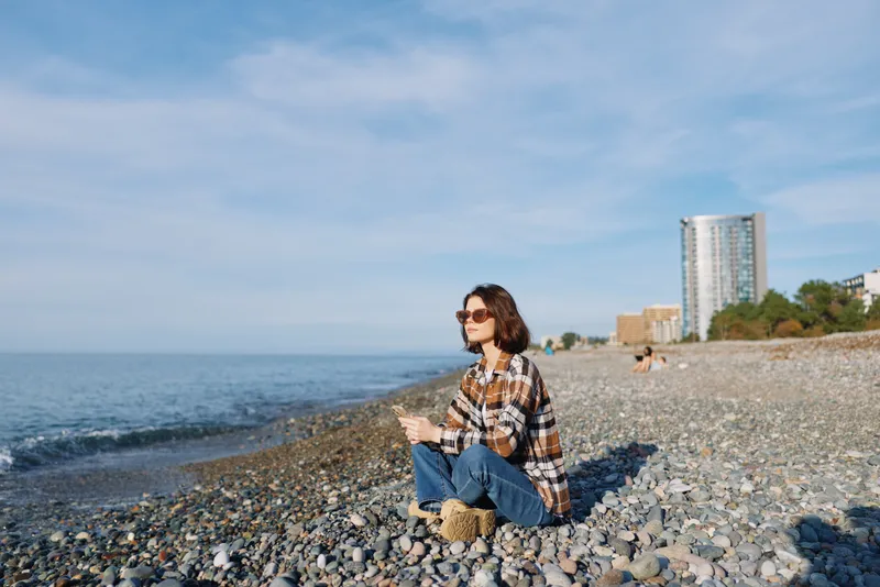 Beach scene with a woman and child sitting on pebbles along the coast, enjoying the calm ocean view, relaxed mood, and sunlit scenery by the water under clear blue sky