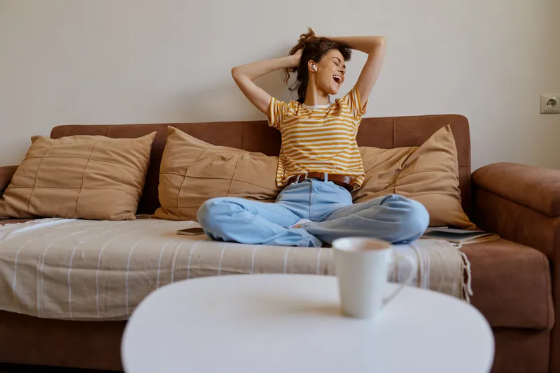 A woman stretches on a cozy couch in a bright living room, wearing a striped yellow and white shirt with blue jeans, exuding a relaxed, casual morning vibe.