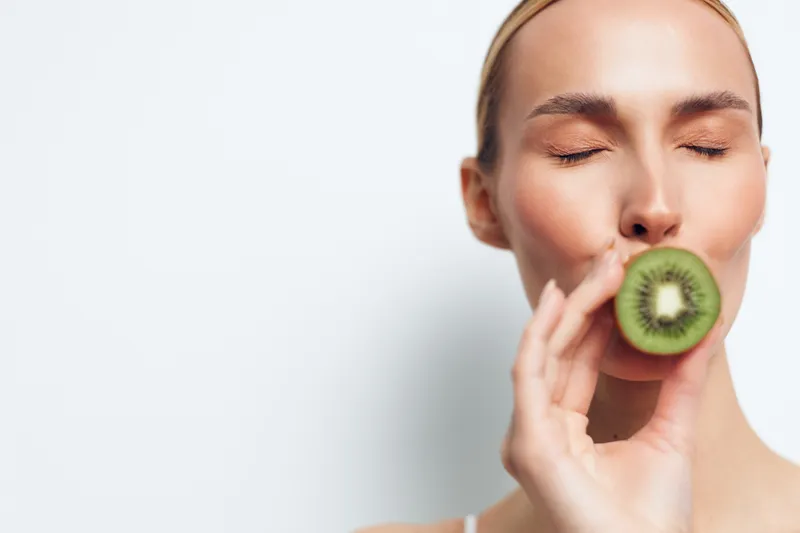 Young woman with smooth skin bites a kiwi slice on her lips, against a clean white background, illustrating fresh fruit skincare, wellness and a healthy lifestyle.