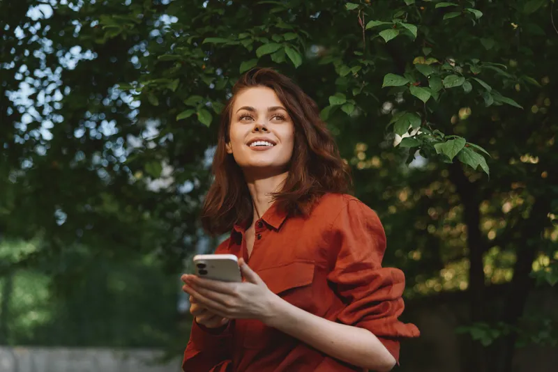 Smiling woman with brown hair wearing a red shirt holds a smartphone outdoors, surrounded by lush greenery. Clear focus on lifestyle moments and natural light