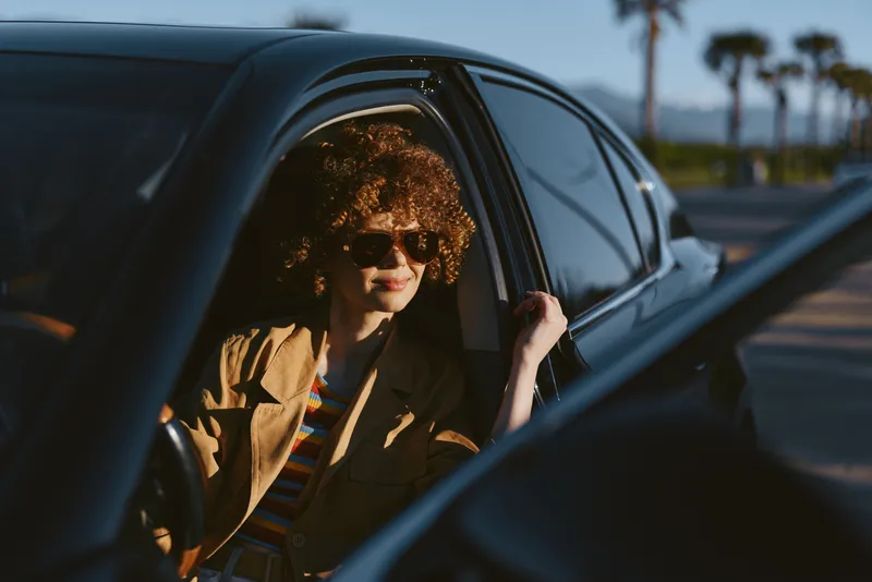 A stylish woman with curly hair in sunglasses sits in a vintage car, sunlit street ahead, embodying carefree road trip vibes and a retro lifestyle moment.