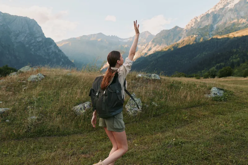 Young woman hiking outdoors in mountains, wearing backpack and casual clothes, raising hand, enjoying nature, sunny day, green grass and rocky peaks background