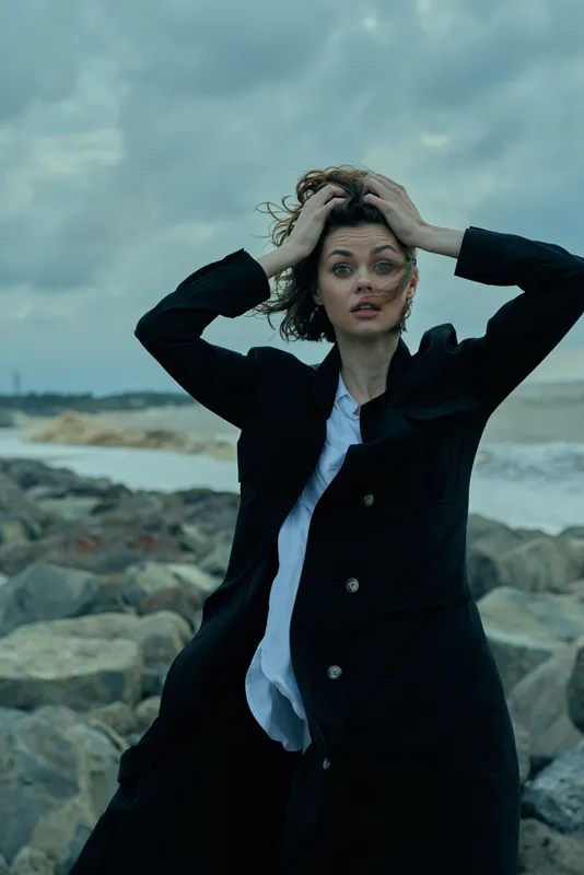 Woman on a windy coast wearing a long dark coat, hands in her hair, intense expression against a rocky seashore, moody lighting, outdoor fashion mood and dramatic atmosphere.