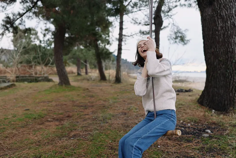 Woman enjoying a rope swing in an outdoor park, laughter and nature mood, casual sweater and jeans, trees and open space framing a carefree moment