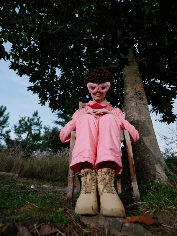 Child in pink outfit sits on a wooden chair under a tree outdoors, wearing chunky boots, captured in a candid moment amid nature and rustic surroundings, the scene emphasizing simplicity and