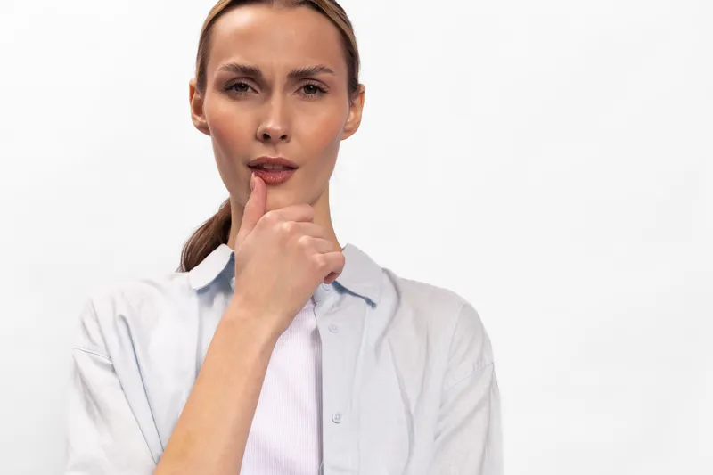 Thoughtful woman with light skin and tied back hair looks at camera with finger on lips, wearing light shirt. Concept for silence, contemplation, and decision making.