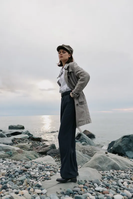 Young woman in trench coat and cap stands on rocky beach near calm sea under cloudy sky. Casual fashion outfit with wide pants creates moody outdoor lifestyle concept by water.