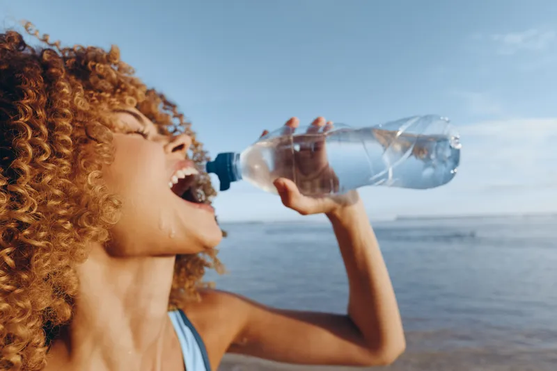 Cheerful woman with curly red hair outdoors by the sea, drinking clear water from a plastic bottle, summer sunlight, relaxed pose, blue sky and calm waves, fresh hydration and wellbeing moment