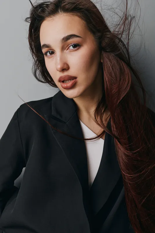 Portrait of young woman with long hair wearing black blazer and white shirt looking at camera with natural makeup and loose hairstyle against neutral background in studio.