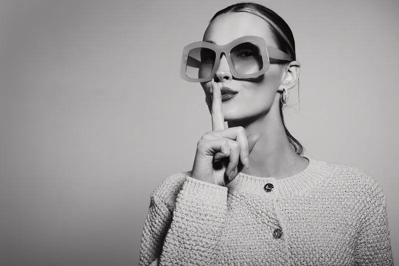 Elegant white woman with pale skin wears oversized sunglasses and knit sweater, holding finger to lips in a quiet gesture.