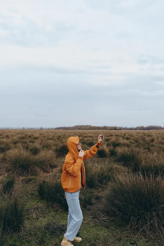 Woman outdoors in grassland field, orange jacket and blue jeans, holding a smartphone, walking and posing, cloudy sky, soft natural light, lifestyle moment, open landscape, quiet trip mood