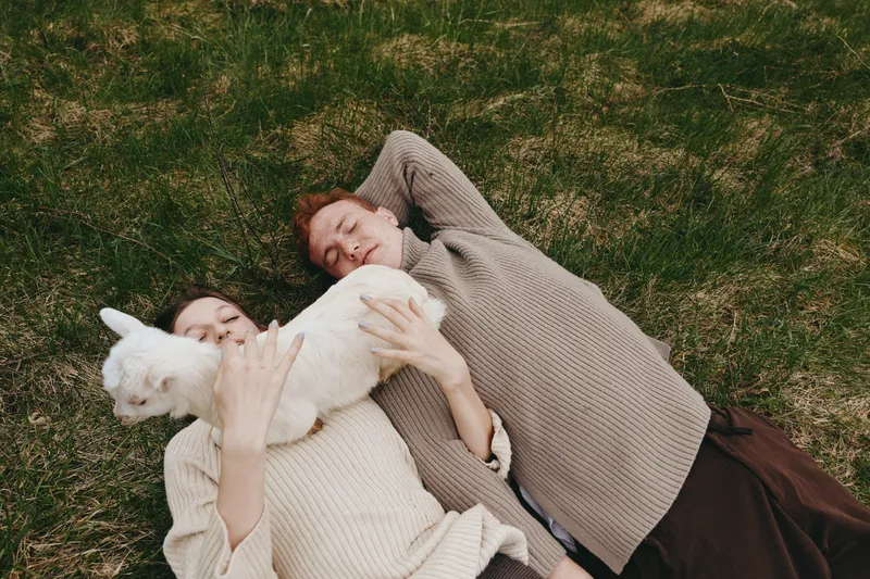 Casual outdoor moment with two friends and a playful goat kid lying on grass, cozy sweaters, soft light, relaxed smiles, natural colors, rustic atmosphere, lifestyle scene outdoors