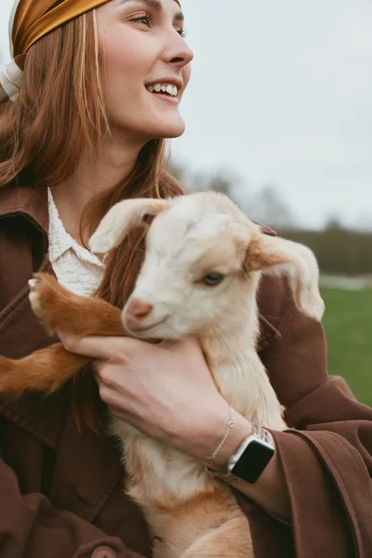 A smiling woman in a brown jacket holds a young goat kid in an outdoors field, capturing a warm lifestyle moment with animals. A gentle bond between human and livestock reflects rural life and care.