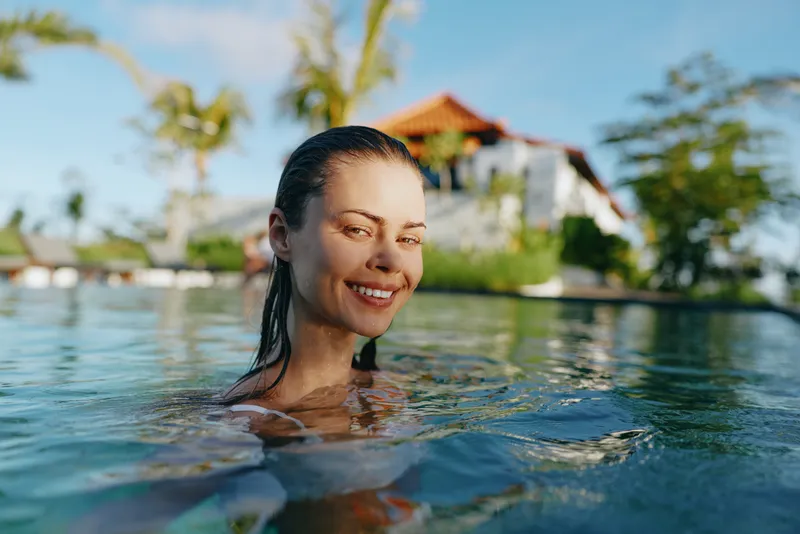 Cheerful woman outdoors, swimming in a sunlit pool, smiling with eyes closed, clear water and palms in the background, relaxed posture, vibrant colors, vacation mood, light breeze, casual swim