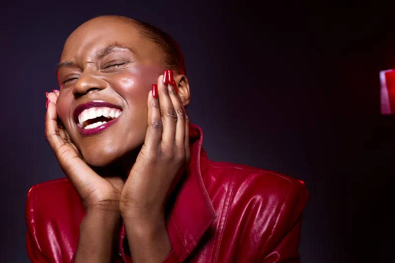 Joyful dark skinned African woman with closed eyes laughs with hands near face wearing red leather jacket. Studio portrait with expressive emotion and vibrant mood on dark background.