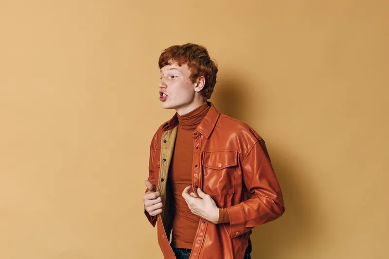 Young man with red hair wearing a brown turtleneck and leather jacket posing against beige background, showing expression and casual fashion style with attention to detail.