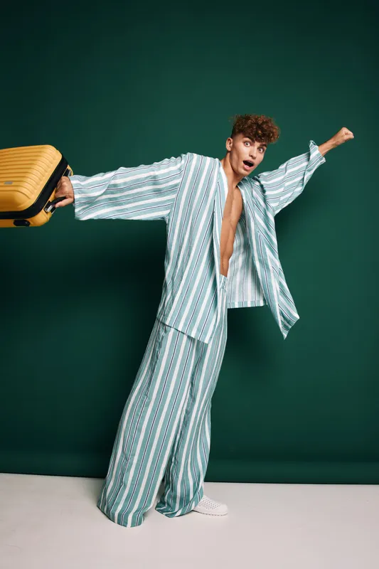 Young man with striped pajamas standing on one leg carrying yellow luggage against dark green background, casual style and travel sign with dynamic motion and cheerful attitude.