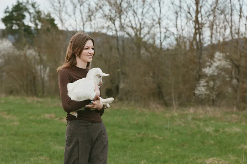 Cheerful woman outdoors, holding a goat kid, gentle smile, casual sweater, sunlit meadow, soft breeze, tranquil moment, rustic vibe, natural colors, countryside reassurance in a peaceful scene