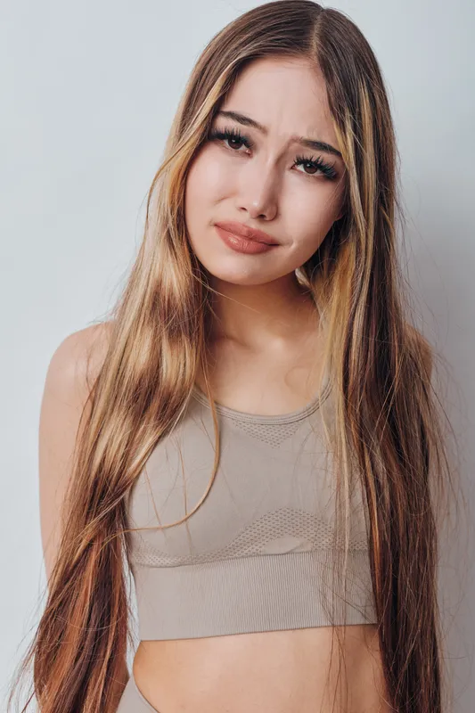 Young woman with long brown hair wearing a beige crop top posing against white background with confused and uncertain facial expression looking at camera.