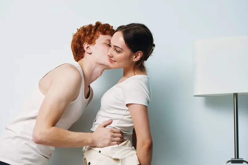 Young couple with white shirts in romantic moment hugging and kissing near lamp in apartment interior for love and affection.