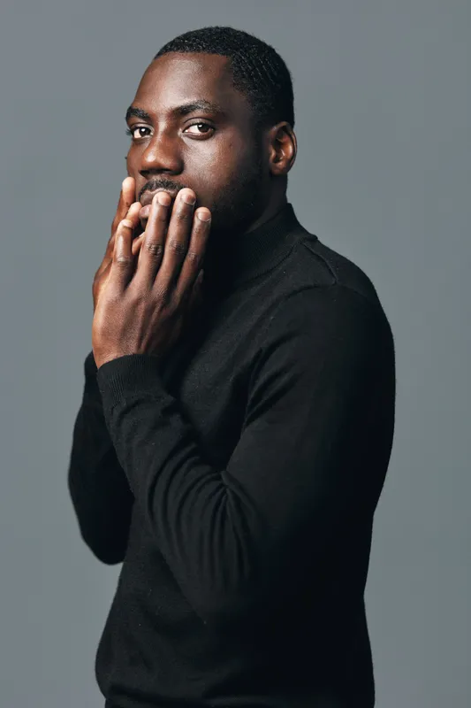 African man with dark skin wearing black sweater in creative conceptual visual style posing with hands near mouth on gray background. Portrait with thoughtful expression and studio lighting.