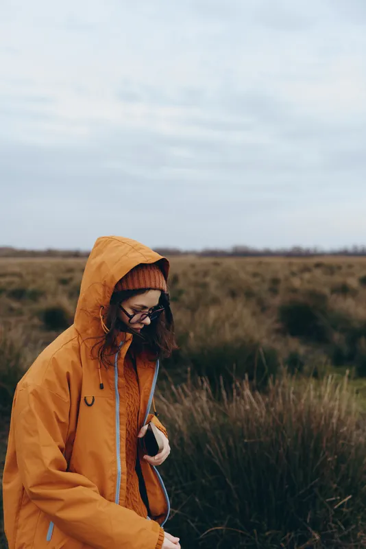 Woman lifestyle outdoors in countryside field wearing yellow raincoat hood and face mask holding smartphone in overcast light cautious mood protective daily life