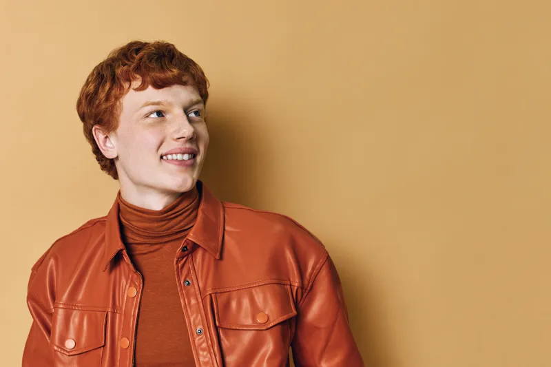 Redhead man with smile wearing brown leather jacket and turtleneck looking sideways on beige background. Portrait, fashion, casual style with warm colors and relaxed mood.