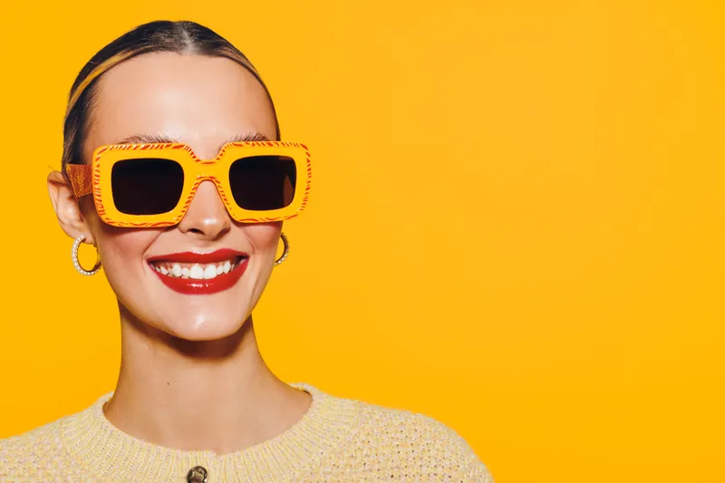 Smiling woman with bright orange sunglasses and red lipstick poses against vibrant yellow background. Concept of summer fashion, happiness, and stylish accessories for modern lifestyle.