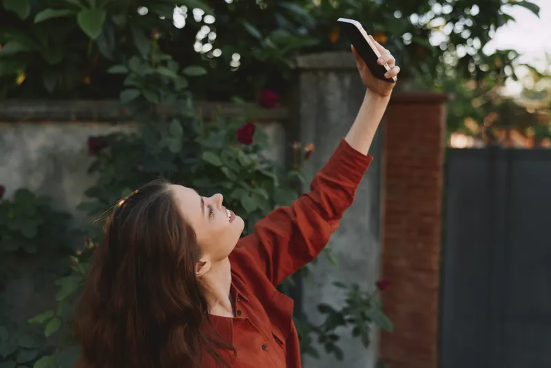 Young woman with long brunette hair lifts a phone to capture a sunny garden moment, wearing a red blouse with natural light and lush greenery in the background