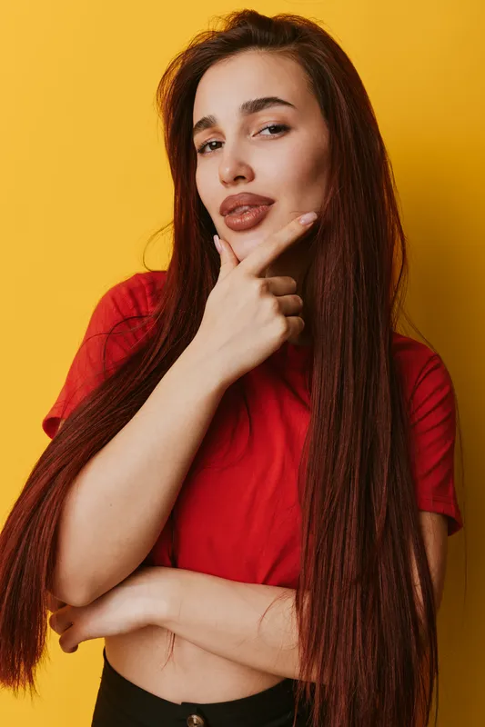 Young woman with long brown hair and red shirt posing with hand on chin thoughtful and confident expression on yellow background. Portrait of casual style and beauty.