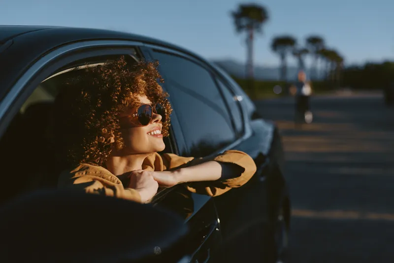 Woman enjoys a sunny drive, leaning from a car window with sunglasses, smiling, wind in her hair, warm light, casual style, outdoor vibe, palm trees in the background, retro mood.