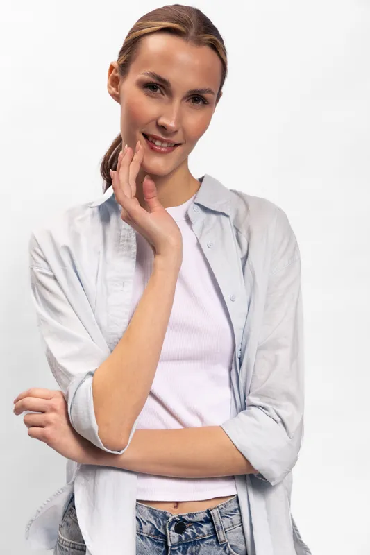 Young woman with light brown hair smiles gently, wearing casual white shirt and jeans. She poses with hand near face on white background. Concept for casual style and natural beauty.