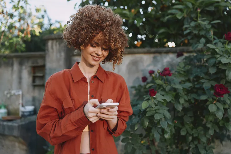 A young woman with curly hair smiles softly while checking her phone in a sunlit garden, wearing a rust colored shirt and enjoying a relaxed outdoor moment