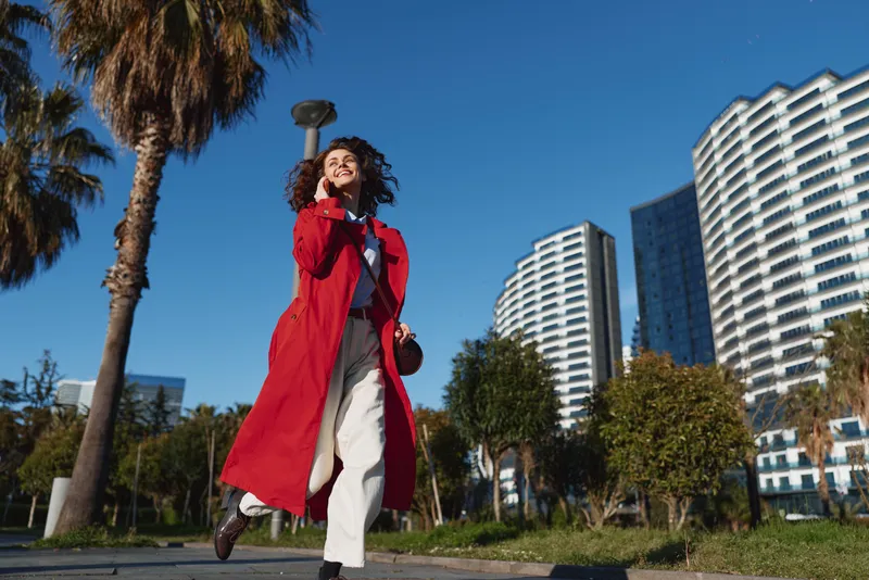 Young woman with curly hair wearing a red coat walks confidently outdoors on a sunny day with modern buildings and palm trees in the background.