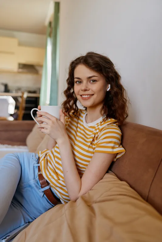 Woman relaxing at home on a sofa, holding a mug, wearing a striped shirt, warm natural light, cozy living space, cheerful everyday lifestyle moment