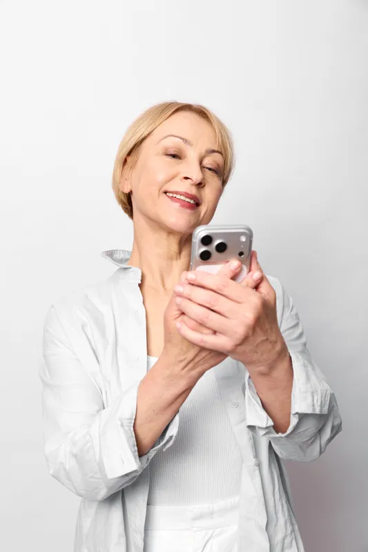 Woman elderly smartphone smiling eyes closed portrait studio technology communication, relaxed hands holding phone, bright white background, soft natural light, casual shirt