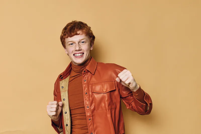 Young man with red hair smiling and posing with raised fists against beige background. Casual style in brown jacket and turtleneck for confident and happy mood.