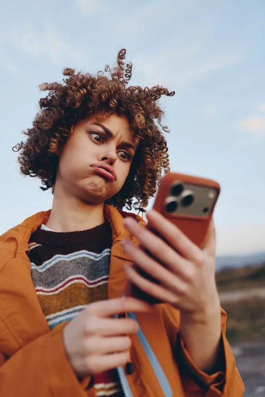 Woman outdoor lifestyle selfie with smartphone, curly hair blowing lightly, orange jacket and striped sweater, holding phone in foreground, scenic sky and distant landscape, portrait