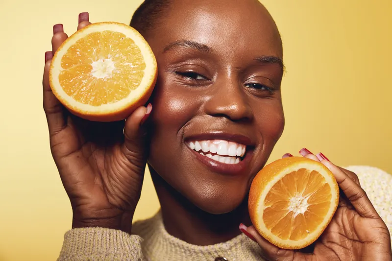 Close-up portrait of a smiling dark skinned African woman holding two halves of fresh orange near her face. Concept of healthy lifestyle, natural beauty, vitamin C, and skincare wellness.
