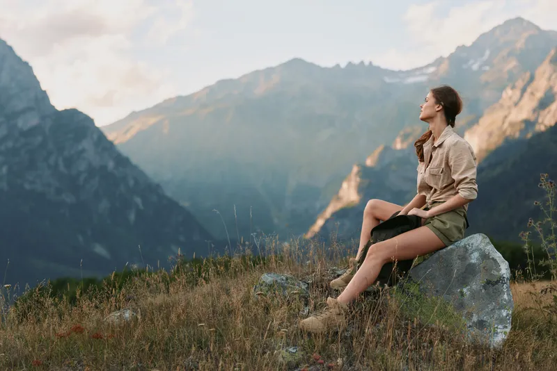 Young woman with long hair sits on a rock in the mountains enjoying the peaceful natural landscape during a calm day.