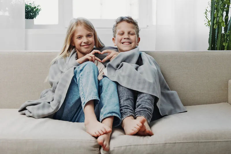 Two happy children wrapped in a cozy gray blanket sitting barefoot on a beige sofa in a bright living room. Concept of sibling love, warmth, relaxation, and family bonding at home.