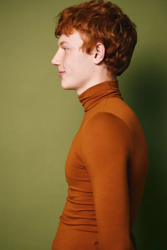 Redhead man in brown turtleneck sweater posing in profile against green background studio. Young caucasian male with curly hair, side view and casual clothing for fashion concept.