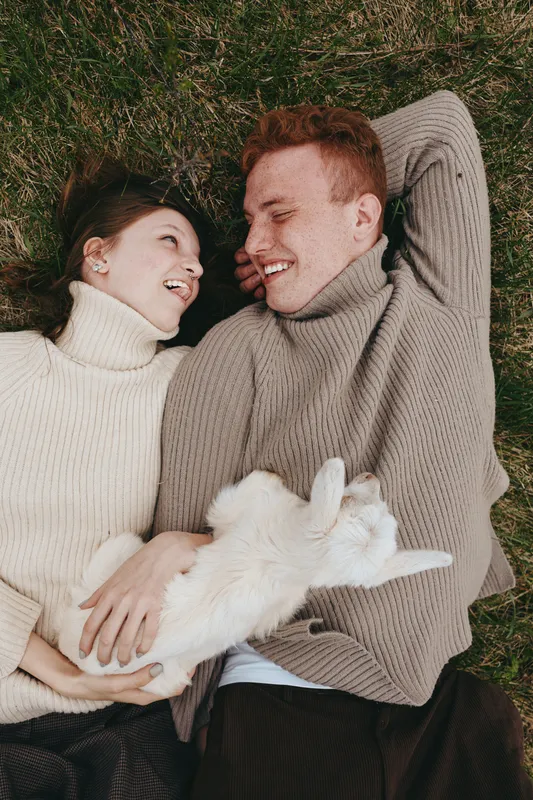 A couple lies on the grass with a playful goat kid, smiling in cozy sweaters. An intimate, relaxed lifestyle moment outdoors, showing affection and a gentle bond with a content animal companion.