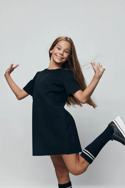 Girl in black dress posing with raised arms and one leg bent on grey background. Smiling child with long hair in casual outfit and sporty socks for youth fashion and joy.